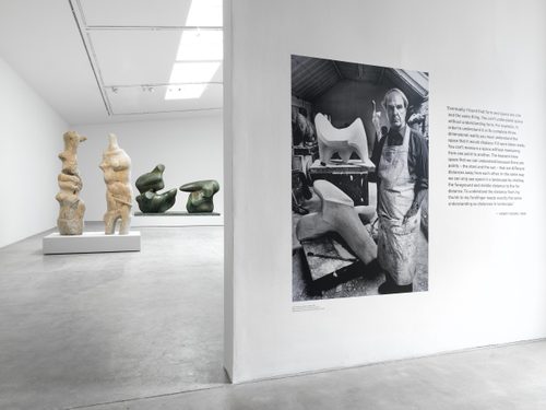 A vintage black and white photo of Henry Moore working in his studio, printed at life size and displayed on a gallery wall. There is a quote by Moore next to it, and a couple of his sculptures can be seen through an opening in the wall behind.