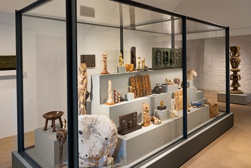 A large display case with a stepped plinth inside, holding numerous small sculptures by Henry Moore made in bronze, plaster and wood. They display a range of styles and techniques, from figurative sculptures to heads and more abstract pieces, and are accompanied by other sentimental objects.