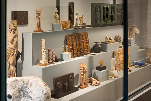 A large display case with a stepped plinth inside, holding numerous small sculptures by Henry Moore made in bronze, plaster and wood. They display a range of styles and techniques, from figurative sculptures to heads and more abstract pieces, and are accompanied by other sentimental objects.