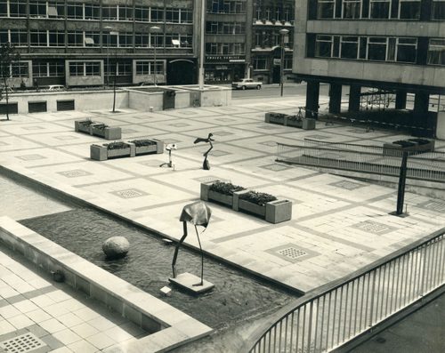 Black and white photo of a large, paved public space between multi-storey buildings. It has a water feature, in the middle of which stands an umbrella-like sculpture. Two more sculptures can be seen further back in the middle of the paved area.