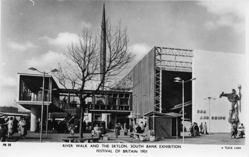 Black and white photo of a large outdoor exhibition space filled with people. A sculpture of a man holding a spear and fish are on the side of a building that reads 'sea and ships'.