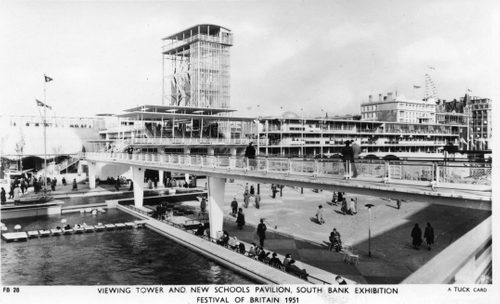 Black and white photo of a pedestrian bridge by a large artificial lake, with a viewing tower behind.