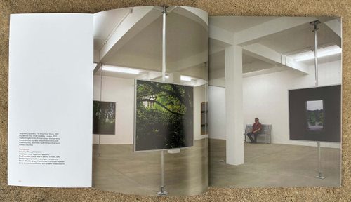 A book lies open on a brown surface on a page with a large photograph of a gallery installation which bleeds over two pages and some acknowledgements on the left.
