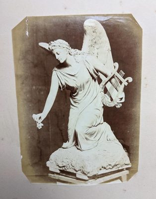 Black and white photo of a sculpture of an angel holding a harp. She is bending down to offer a flower to some unseen person.