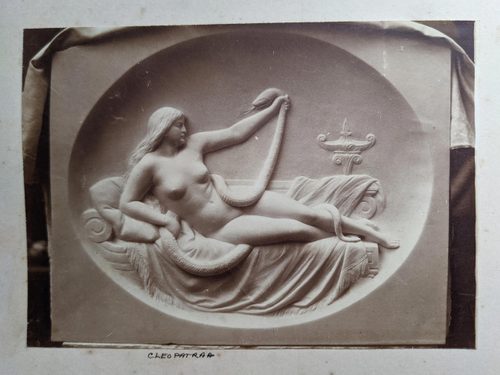 Black and white photo of a relief sculpture of a naked woman lying on a chaise longue, gripping the head of a snake in one hand, her arm outstretched to keep it at a distance from her body.