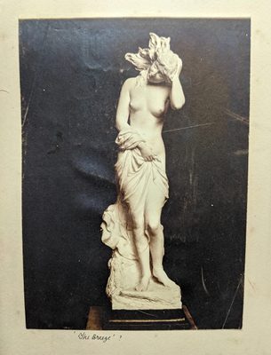 Black and white photo of a sculpture of a naked young woman. She clutches a sheet to her waist, and her hair is blowing in a breeze.