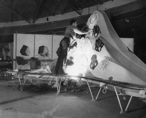 Black and white photo of a man standing on a rail to work on a dome-shaped sculpture, which is covered with drawings of insects, each around two feet long.