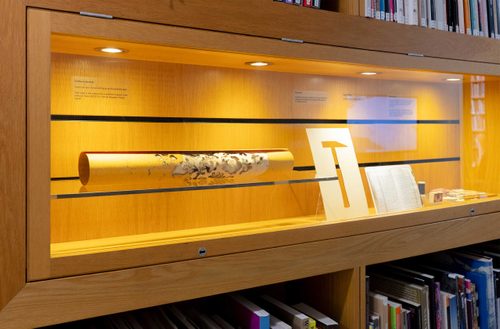 A display cabinet with a rolled up drawing, stencil of a hammer, open book, and stationary.