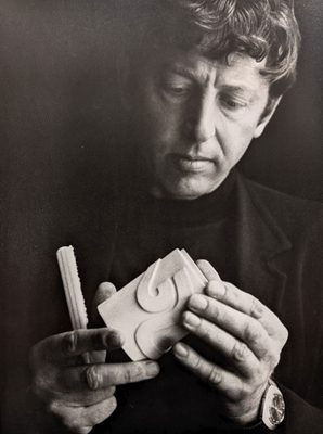 Headshot of Geoffrey Clarke, who is holding a small sculpture and brush. He is dressed in smart black clothes and is wearing a watch.