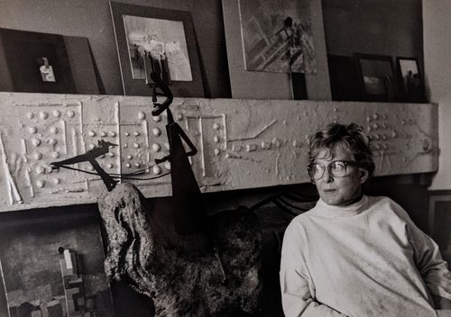 Black and white photo of an older woman with large glasses looking to her right, where there is an abstract sculpture of a horse and rider made from what looks like metal and stone. Behind her are more sculptures and paintings.