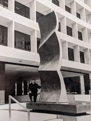 Black and white photo of a man stood next to a large piece of public sculpture outdoors. The sculpture is maybe 20 to 25 feet tall but quite thin, and shaped like a wave.