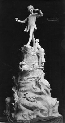 Black and white photo of a sculpture of Peter Pan, who is playing a flute while stood on a hill, which is decorated with rabbits and fairies.