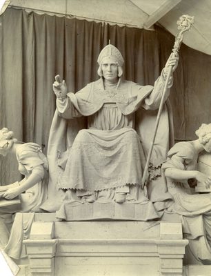Black and white photo of a sculpture of a seated man, wearing robes and mitre that indicate he is of the Christian faith. He is making a blessing sign with his right hand and holding a staff with a bell in his left. Two women, also part of the sculpture, are seated at his feat, reading.
