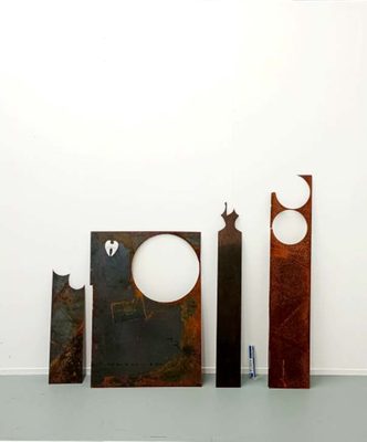 Four sheets of rusted metal with circles cut out of them, leaning against a while gallery wall.
