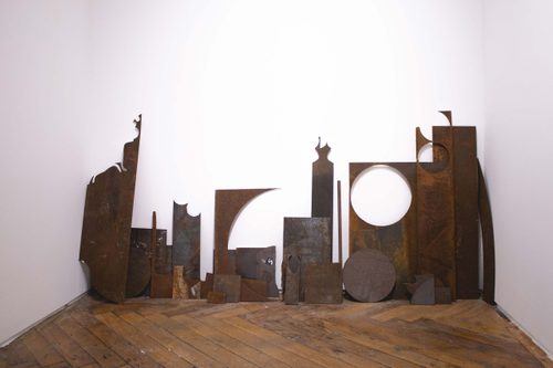 Several sheets of rusted metal with circles cut out of them, leaning against a while gallery wall.