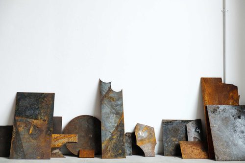 Several sheets of rusted metal with circles cut out of them, leaning against a while gallery wall.