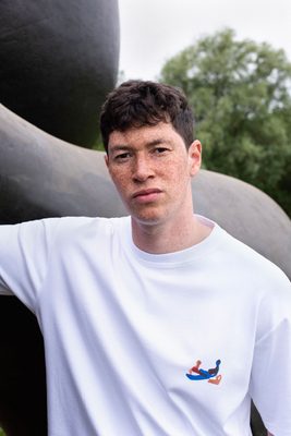 A white man with a freckled face and brown hair faces away from the camera. He is wearing a white t-shirt with a small abstract design by Henry Moore on the left breast, depicting a reclining figure drawn in blue, brown and red felt-tip pen. Behind the model is a bronze sculpture by Henry Moore.