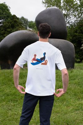 A white man with a freckled face and brown hair faces away from the camera. He is wearing a white t-shirt with an abstract design by Henry Moore of a reclining figure drawn in blue, brown and red felt-tip pen. Behind the model is a bronze sculpture by Henry Moore.
