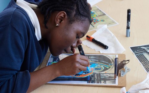 A young girl drawing bold colours over the top of a photo of a sculpture on a clipboard.