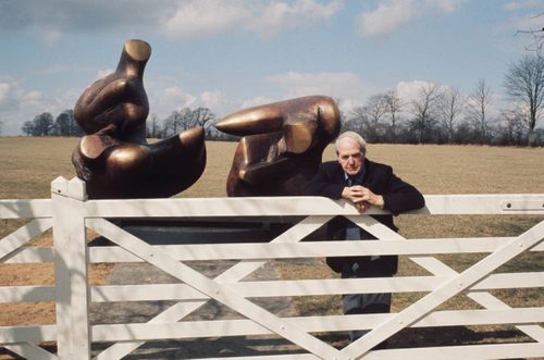 A colour photo of a older man leaning on a five-bar gate. Behind him is a large sculpture made of bronze.