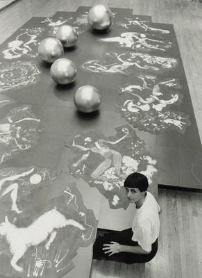 Black and white photo of a woman sat on the floor making a large-scale artwork, featuring drawings of a sheep, a many-armed figure, and many others, spread out among five large metallic spheres.