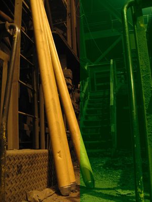 Photograph of an industrial space, with three long metal tubes leaning against a wall and metal stairs behind. The photo has been coloured yellow on the left half and green on the right half.