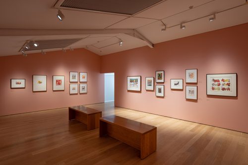 Installation view of a gallery, with 15 framed drawings and prints hanging on two walls which meet in a corner. The walls are painted salmon pink and the lighting is dimmed. All of the artworks are abstract and semi-abstract works by Henry Moore.