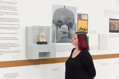 A woman with dyed pink hair wearing black stands to read part of a timeline displayed on the wall of the Henry Moore: The Sixties exhibition. Mounted in a small display case is a plaster maquette for a sculpture by Henry Moore.