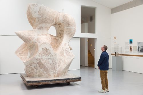 A young man wearing a blue jacket and tan trousers stands to contemplate a huge plaster sculpture by Henry Moore. They are located in a large gallery space with white walls and a pale grey floor.