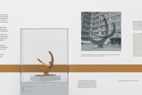 Part of the timeline in the Henry Moore: The Sixties exhibition. A large black and white photo on the wall shows Henry Moore's sculpture 'Sundial' in the forecourt of the Times newspaper offices. Mounted in a small display case next to the photo is a small model of the sculpture.