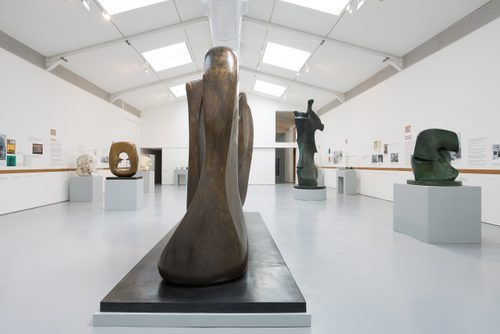 A wide view of the Henry Moore: The Sixties exhibition, a huge bronze sculpture 'Kinfe Edge: Two Piece' looms close to the camera, with four other large sculptures arranged in the background. The gallery walls are white and the floor is grey. There are skylights in the roof.