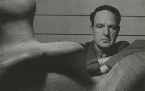A black and white photo of Henry Moore, a man with dark hair in his 40s, standing behind an abstract, carved, wooden sculpture of a female human form.