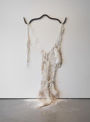 Abstract sculpture made from white kaolin clay coating coarse raffia fibres, hanging from an iron gambrel attached to a wall.