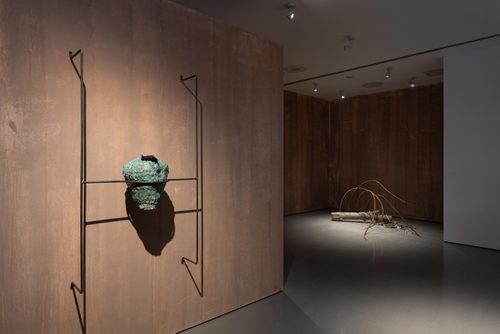 A wire armature mounted to a wall displays a sculpture of roughly-made pot, painted with a copper-green patina. On the floor behind is a sculpture made from a driftwood log, with four long, rusted metal spikes reaching out like a breaking wave.