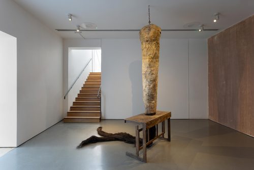 A sculpture hangs from a chain in a gallery. The upper part is roughly cylindrical and yellow-cream coloured. It descends through a hole cut in an old wooden desks, and where it emerges it is made of hair-like fibres, which rest on the floor in two 'tails'.