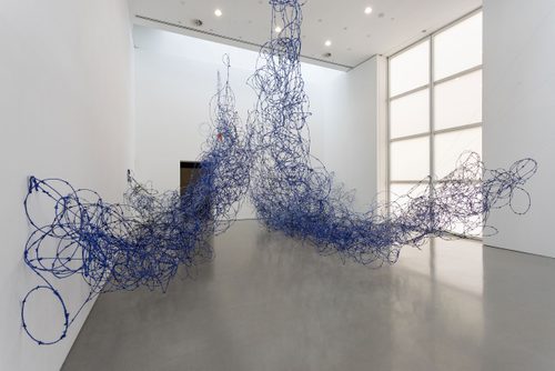 Photograph of the exhibition 'Lungiswa Gqunta: Sleep in Witness', showing the artist's sculpture 'Ntabamanzi' made from barbed wire and blue wool. The sculpture is designed to look like a wave breaking against the wall of the gallery.
