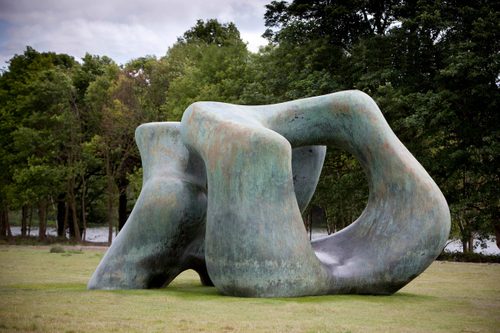 Large green-tinged bronze sculpture in two parts, with a hole through the middle. It is in a field of grass with trees behind.