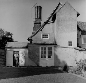 A black and white photo showing the rear of an old farmhouse. A man, in his early 40s, is standing in an open doorway in the left of frame.