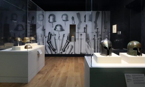 Display cases filled with helmets from the Wallace Collection, from simple bronze helmets made in Ancient Greece to helmets used in World War I and II. A large photo of various medieval arms and armour is on the wall behind.
