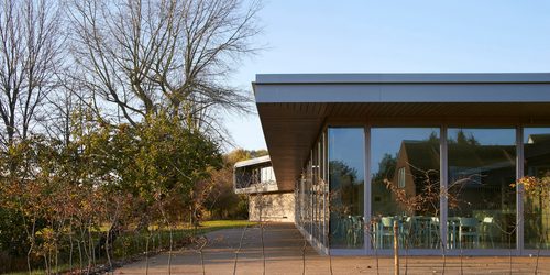A photo of contemporary styled café building, taken from outside. Reflected in the windows is a character cottage.