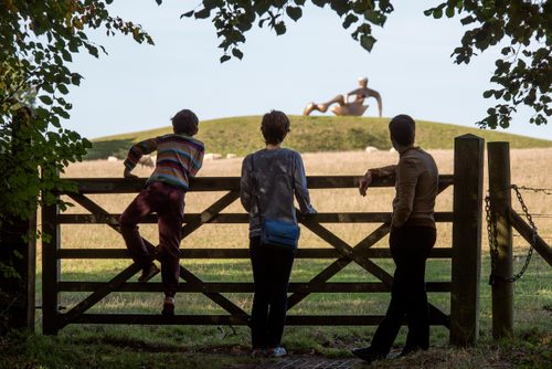 A family of three looking over a five-bar gate at Henry Moore's sculpture Large Reclining Figure, which sits on a hill in the distance