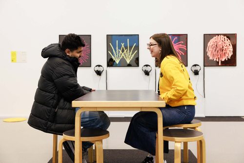 An exhibition guide in a bright yellow 'Beyond the Visual' top is sat with a visitor at a table in a gallery. Mounted on the wall behind them are four artworks that look like graphical depictions of fireworks exploding.