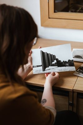Photo looking over the shoulder of a young woman with brown hair and a small tattoo on her right forearm. She is holding a black and white photo of an large geometric sculpture set in the landscape. More photos are on the desk in front of her.