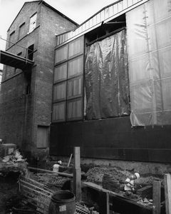 Black and white photo of a building under construction. A wide, two-storey opening has been made in one wall, and is covered by plastic sheeting. Two workmen are laying bricks in the street below, possibly for foundations or for a basement level.