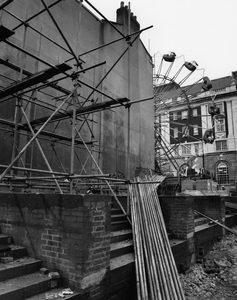 Black and white photo of a building under construction, with steps leading up to a blank wall with scaffolding being set up. A Ferris Wheel can be seen in the background.