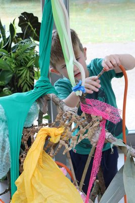 A boy wearing a blue/green t-shirt using sticks, fabric and reclaimed packaging materials to build a sculpture.