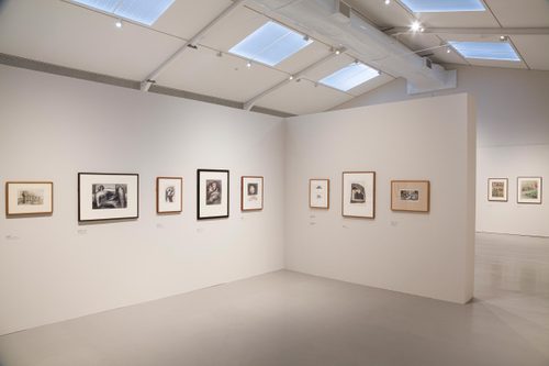 A high-ceilinged gallery space with a free-standing wall in the centre, on which several framed drawings are hung.
