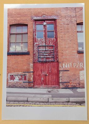 Printed photo of an industrial-looking red brick wall, with large, red wooden double doors, with the paint flaking off. A sign has been mounted to the door, which reads: 