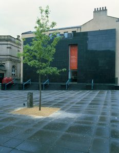 A green sapling with a small upright basalt stone stood next to it, in a paved square in front of a building with an imposing black granite facade.