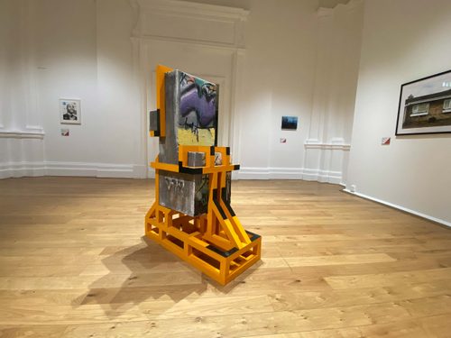 An abstract sculpture made of a rectangular concrete block, covered in graffiti, held in place and wrapped around by a yellow-painted wooden frame.